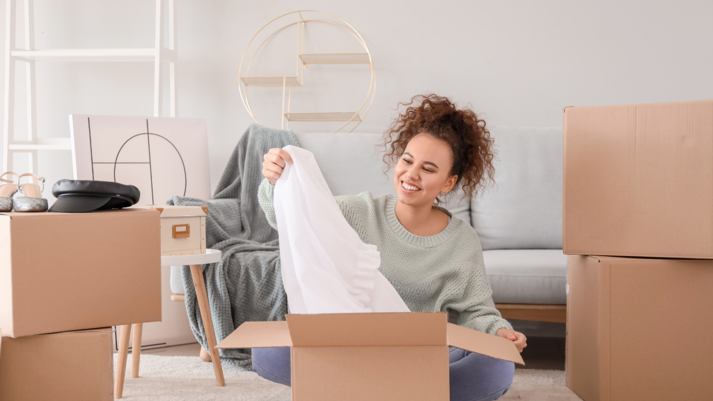 Woman sitting on the floor among boxes, smiling as she pulls a white blouse from an open box.